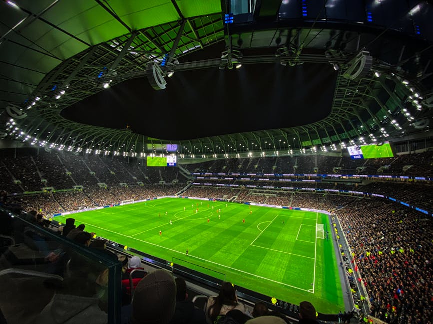 Interior view of a large football stadium during a match, with a well-maintained bright green grass pitch marked with white lines. The stadium is filled with spectators seated in dark tiered stands th